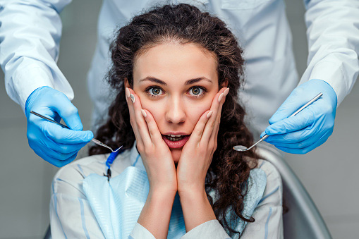 A nervous female patient holding her face while gloved hands approach with dental tools